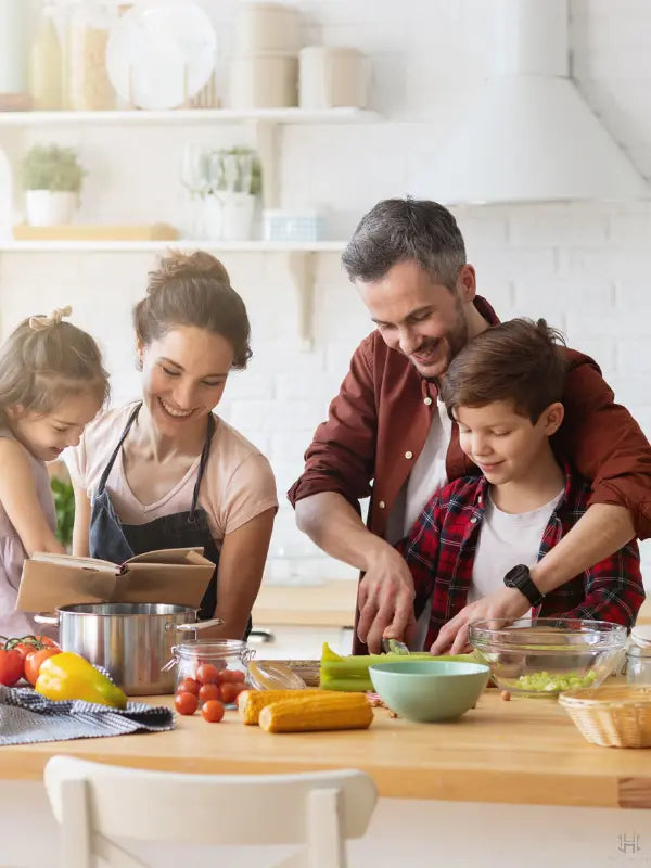A family of four enjoys cooking together in a bright, modern kitchen. The mother reads a book to her two children, who are helping her prepare vegetables on a wooden table.